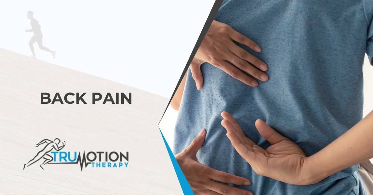 A man with back pain reaching at his lower back with a chiropractor's hands near them | Conditions We Treat | Back Pain | TruMotion Therapy